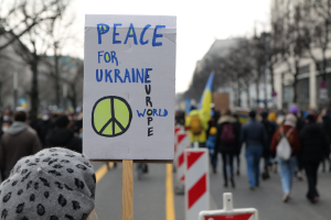 Eine Person mit Mütze hält ein "Frieden für die Ukraine"-Schild vor einer Menge, die auf einer Straße geht, einige halten Flaggen, mit Bäumen, Gebäuden und einem klaren blauen Himmel im Hintergrund.