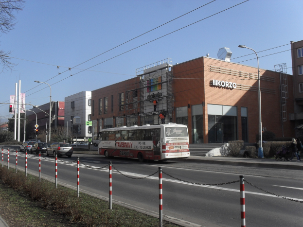 Ein Bus fährt auf einer Straße mit hohen Gebäuden, Straßenlaternen, Strommasten, Stromkabeln, Kraftfahrzeugen, Pollern, Fußgängern auf dem Gehweg, Büschen, Bäumen und einem klaren blauen Himmel.