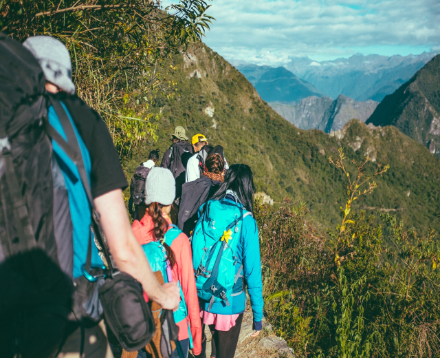 Gruppe von Menschen, die einen Berg hinaufwandern, mit Rucksäcken und Hüten, mit bewölktem Himmel und Bäumen im Hintergrund.