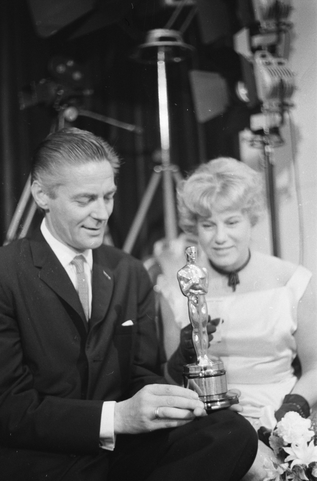 Ein Mann in einem schwarzen Blazer, weißem Hemd und Krawatte hält eine Oscar-Statue, während er neben einer Frau in Abendgarderobe sitzt, die einen Blumenstrauß hält, mit sichtbaren Lichtern im Hintergrund, alles in Schwarz-Weiß.