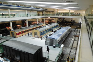 Ein großer Museumsraum mit verschiedenen Bahnen in unterschiedlichen Größen und Farben, mit Menschen, Geländern, Säulen und Deckenlampen.