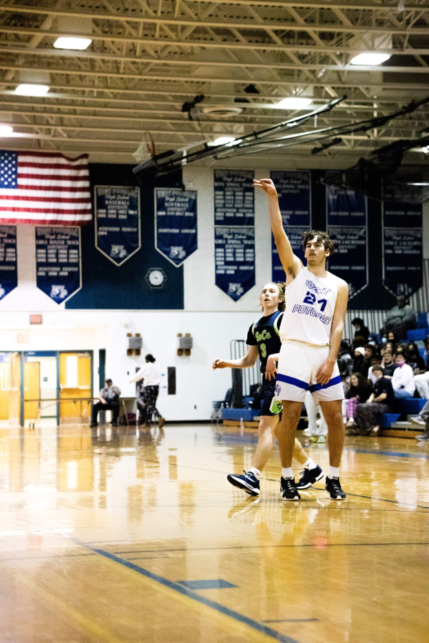 Ein Mann in Sportschuhen schießt einen Basketball in einer Turnhalle, während ihn eine Menge Zuschauer von den Hintergrundbänken und einer Wand mit Bannern und einer Fahne aus anfeuert.