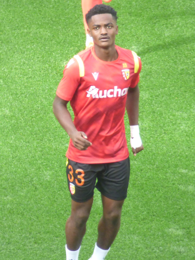 Emmanuel Adebayor in einem roten Trikot und schwarzen Shorts auf einem Fuballfeld mit zwei Personen im Hintergrund.