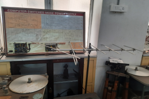 Ein überfüllter Elektronikraum mit einer beschrifteten Leiterplatte auf einem Tisch, Metallstangen, einer wandmontierten Schalttafel, einem Fenster und Gegenständen in Schränken, ähnlich einer computerge-switchten Modus-Stromversorgung.