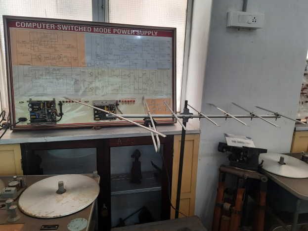 Ein überfüllter Elektronikraum mit einer beschrifteten Leiterplatte auf einem Tisch, Metallstangen, einer wandmontierten Schalttafel, einem Fenster und Gegenständen in Schränken, ähnlich einer computerge-switchten Modus-Stromversorgung.