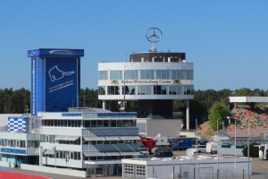 Außensicht der Mercedes-AMG Petronas F1 Team-Zentrale in Berlin, Deutschland, mit Gebäuden, Fahrzeugen, einem Zaun, Beschriftungen, Bäumen und einem Logo auf der Struktur unter einem sichtbaren Himmel.