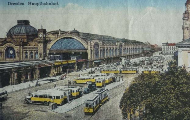 Eine alte Postkarte zeigt einen belebten Bahnhof in Dresden, Deutschland, mit Fahrzeugen, Menschen, Bäumen, Gebäuden und einem bewölkten Himmel, mit der Aufschrift "Dresden Hauptbahnhof" oben.