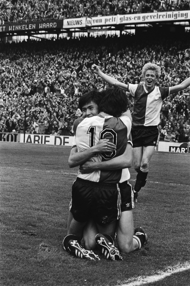 Drei Fußballspieler feiern ein Tor auf dem Boden, umgeben von einer jubelnden Menge und schwenkenden Fahnen im Hintergrund, dargestellt in Schwarz-Weiß.