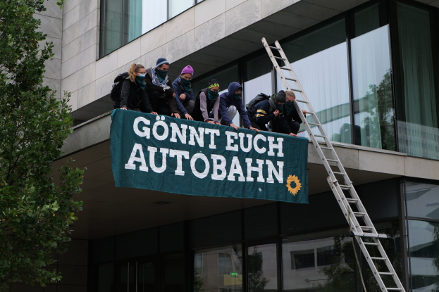 Eine Gruppe von Menschen steht auf dem Dach eines Gebäudes und hält eine Fahne, mit einem Baum links und Glasfenstern im Hintergrund, scheinbar protestierend zur Unterstützung der deutschen Regierung.