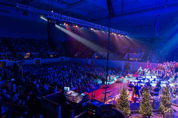 Eine große Menschenmenge in einem geschmückten Auditorium schaut einer Band zu, die auf einer Bühne spielt, die mit Weihnachtsbäumen und festlichen Lichtern geschmückt ist.