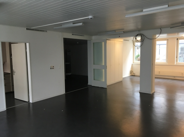 Leeres Zimmer mit schwarzen Böden, weißen Wänden, elektrischen Lampen, Kabeln, Türen, Schränken, Fenstern und einer Aussicht auf Gebäude und Bäume draußen, das sich in einem zu vermietenden Büro in Amsterdam befindet.