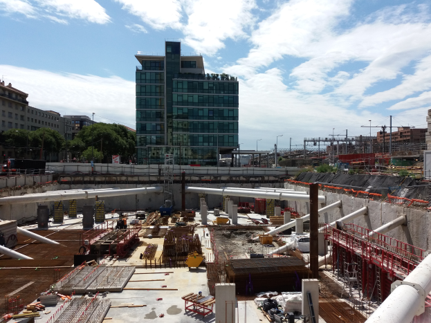 Eine Baustelle mit einem großen Gebäude, wahrscheinlich den europäischen Union-Hauptsitz, umgeben von Bäumen, Pfählen und Lichtern unter einem bewölkten Himmel.