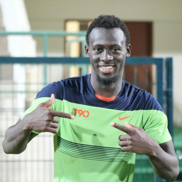 Ein Mann in einem grünen und blauen Hemd, der lachend in die Kamera zeigt und im Hintergrund eine Brüstung, eine Wand und eine Tür zu sehen sind.