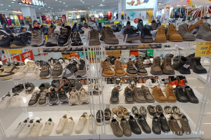 Ein Schuhgeschäftsinneres mit verschiedenen Schuhen auf Gestellen, Preisetiketten sichtbar, Menschen im Hintergrund, Kleiderständer, Deckenlampen, ein Bildschirm und ein Wasserzeichen, das einen Store in China anzeigt.