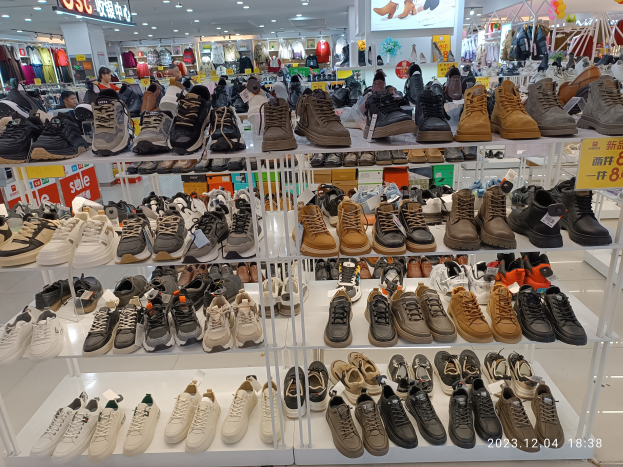 Ein Schuhgeschäftsinneres mit verschiedenen Schuhen auf Gestellen, Preisetiketten sichtbar, Menschen im Hintergrund, Kleiderständer, Deckenlampen, ein Bildschirm und ein Wasserzeichen, das einen Store in China anzeigt.