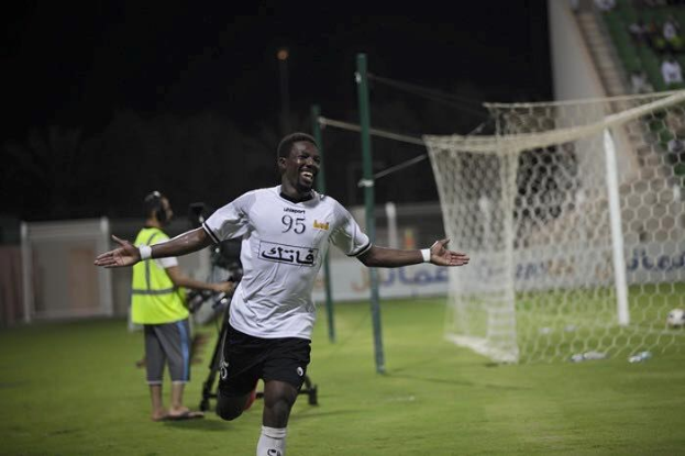 Ein Mann in weißem Hemd und schwarzen Shorts rennt über ein Fußballfeld und breitet die Arme aus, umgeben von Zuschauern, mit einem Tor und sitzenden Zuschauern im Hintergrund.