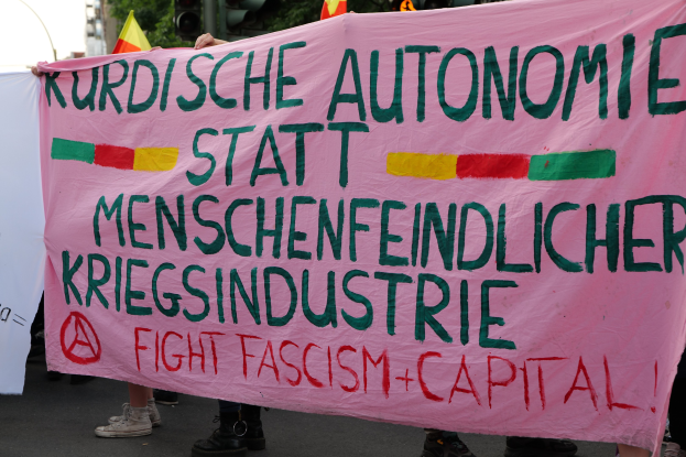 Eine Gruppe von Menschen mit einem pinken Banner, auf dem "Kurdische Autonomie Statt Menschenfeindlicher Kriegsindustrie Fight Fascism Capital" steht, auf einer Straße mit Ampeln, Flaggen, Bäumen, Gebäuden und einem klaren blauen Himmel im Hintergrund.