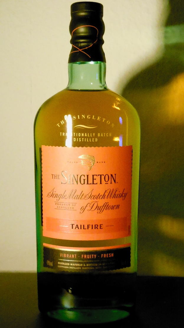 Eine tiefe bernsteinfarbene Flasche Singleton of Dufftown Tailfire Single Malt Scotch Whisky auf einem Tisch, mit einem Etikett, das eine schwarzes Logo und ein Wappen mit einem Löwen und einer Burg zeigt.