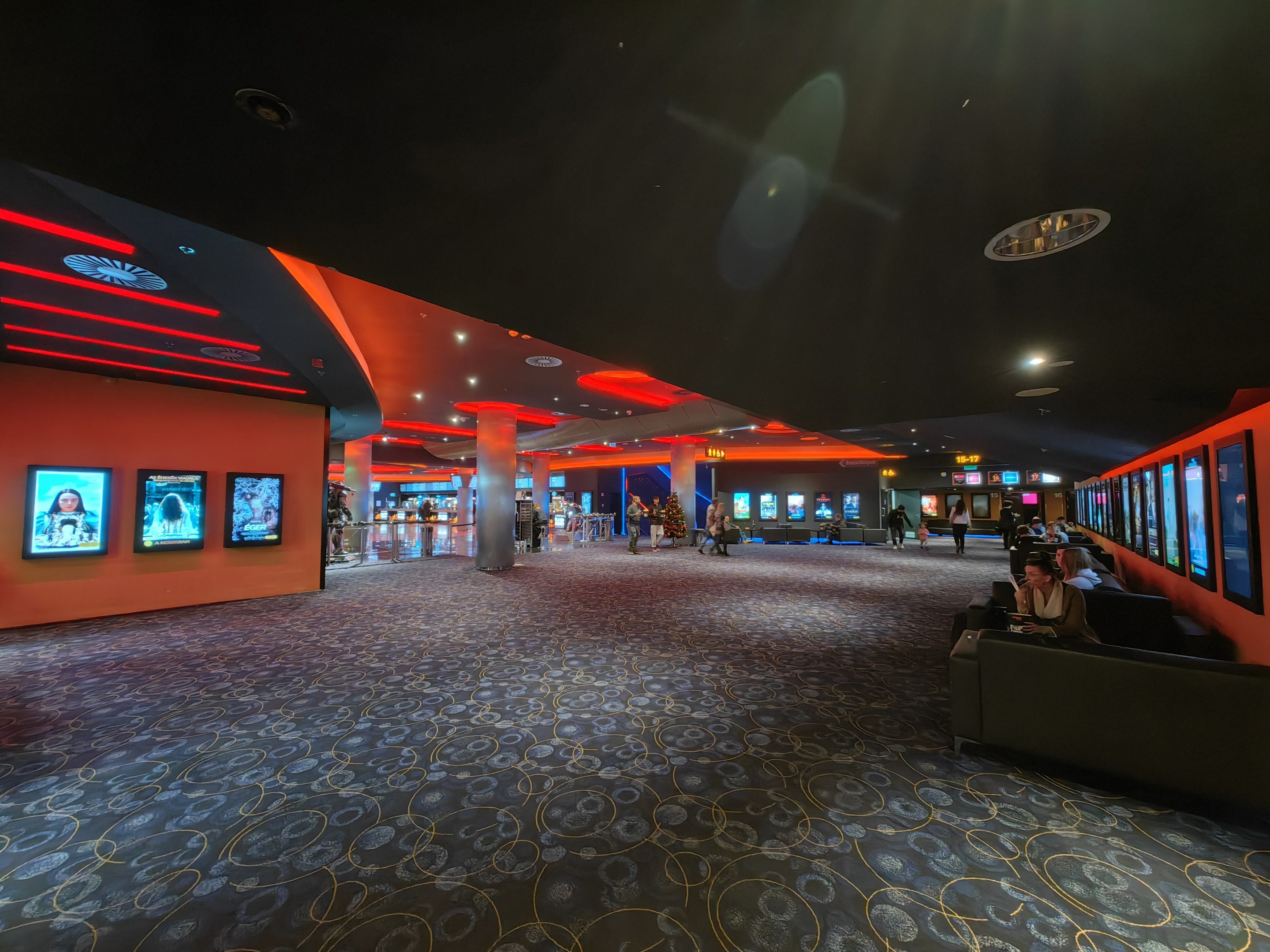 Großer Raum mit Menschen auf Sofas sitzend, die an der Wand montierte Fernseher anschauen, umgeben von Säulen und Deckenleuchten, mit sichtbarem Boden.