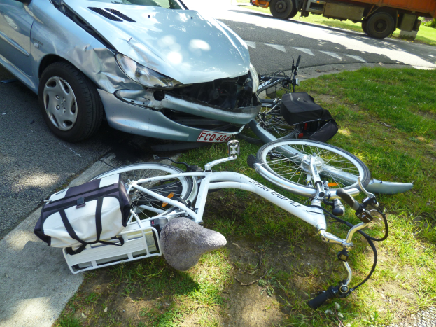Ein verunglücktes Auto am Straßenrand mit einem Fahrrad auf dem Gras daneben, Bäume im Hintergrund und einen klaren blauen Himmel.
