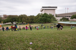 Eine Gruppe von Menschen spielt mit Ostereiern auf einer grünen Wiese mit Bäumen, Gebäuden, Flaggen, einem Brunnen und einem bewölkten Himmel im Hintergrund.