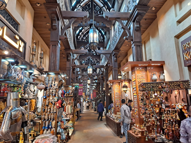 Ein geschäftiges Dubai-Markt mit Menschen zwischen Ständern mit Waren, Wänden mit Texttafeln und einer Decke mit warmem Licht.