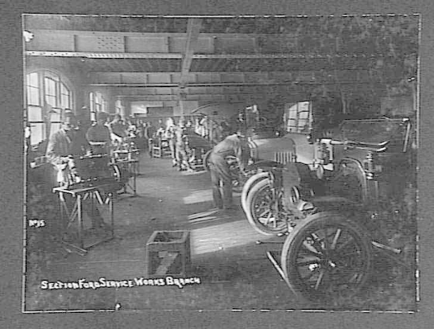 Schwarzes und weißes Foto von Männern, die an einem Auto in einer Ford-Werkstatt arbeiten, mit Fahrzeugen, Werkzeugen und Fenstern im Bild, einschließlich Text unten.