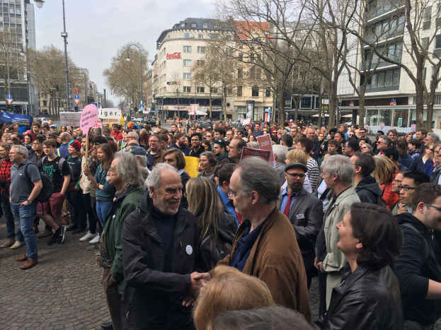 Eine große Gruppe von Menschen, die vor einer Menge mit Schildern steht, mit Fahrzeugen, Bäumen, Laternenpfählen und Gebäuden im Hintergrund, wahrscheinlich während einer Demonstration in Berlin, Deutschland.