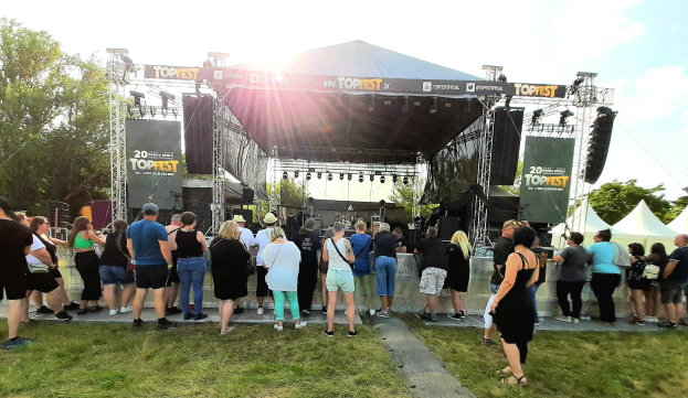 Menschenmenge vor einer Bühne auf einem Musikfestival mit Zelten und Bannern rechts, grasigem Bereich, Bäumen, bewölktem Himmel und strahlendem Sonnenlicht.