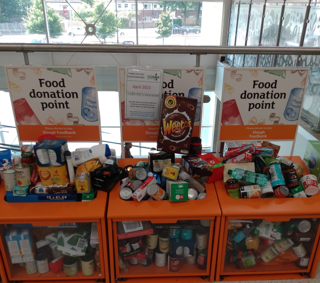 Orange Schränke voller Lebensmittelspenden, darunter Flaschen, Dosen und Kartons, neben Informationsschildern; Hintergrund zeigt Fahrzeuge, Bäume, Gebäude und Himmel durch Glasfenster.