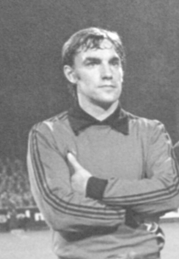Schwarzes und weißes Foto eines Mannes auf einem Fußballfeld mit verschränkten Armen, umgeben von sitzenden Zuschauern im Hintergrund.