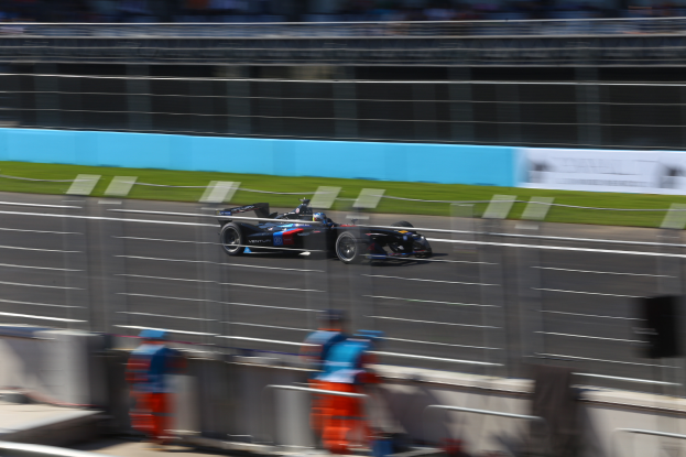Ein Formelwagen fährt auf einer Rennstrecke mit einer unscharfen Zuschauermenge am Rand, Gras am Boden und Tafeln mit Text im Hintergrund.