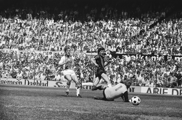Schwarzes und weißes Foto von Männern, die auf einem Feld Fußball spielen, mit Zuschauern im Hintergrund und einer 'Bundesliga 1978-79 Ajax 1-1 Hannover 96'-Fahne.