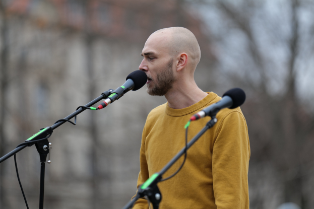 Ein Mann mit Glatze in einem gelben T-Shirt singt in ein Mikrofon mit unscharfem Hintergrund.