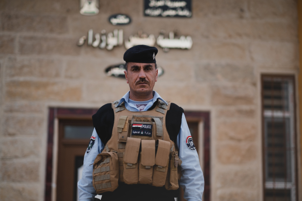 Ein Polizist in Uniform und Mütze steht vor einem Gebäude mit Türen, Fenstern und Text an der Wand.