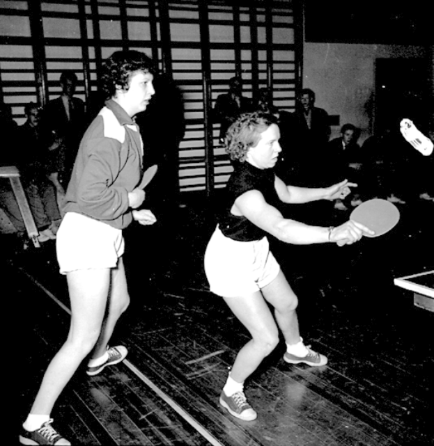Zwei Frauen spielen Tischtennis auf einem Court, jede hält einen Schläger in der Hand, mit einigen Zuschauern im Hintergrund und einem Tischtennis-Tisch auf der rechten Seite, dargestellt in Schwarz-Weiß.