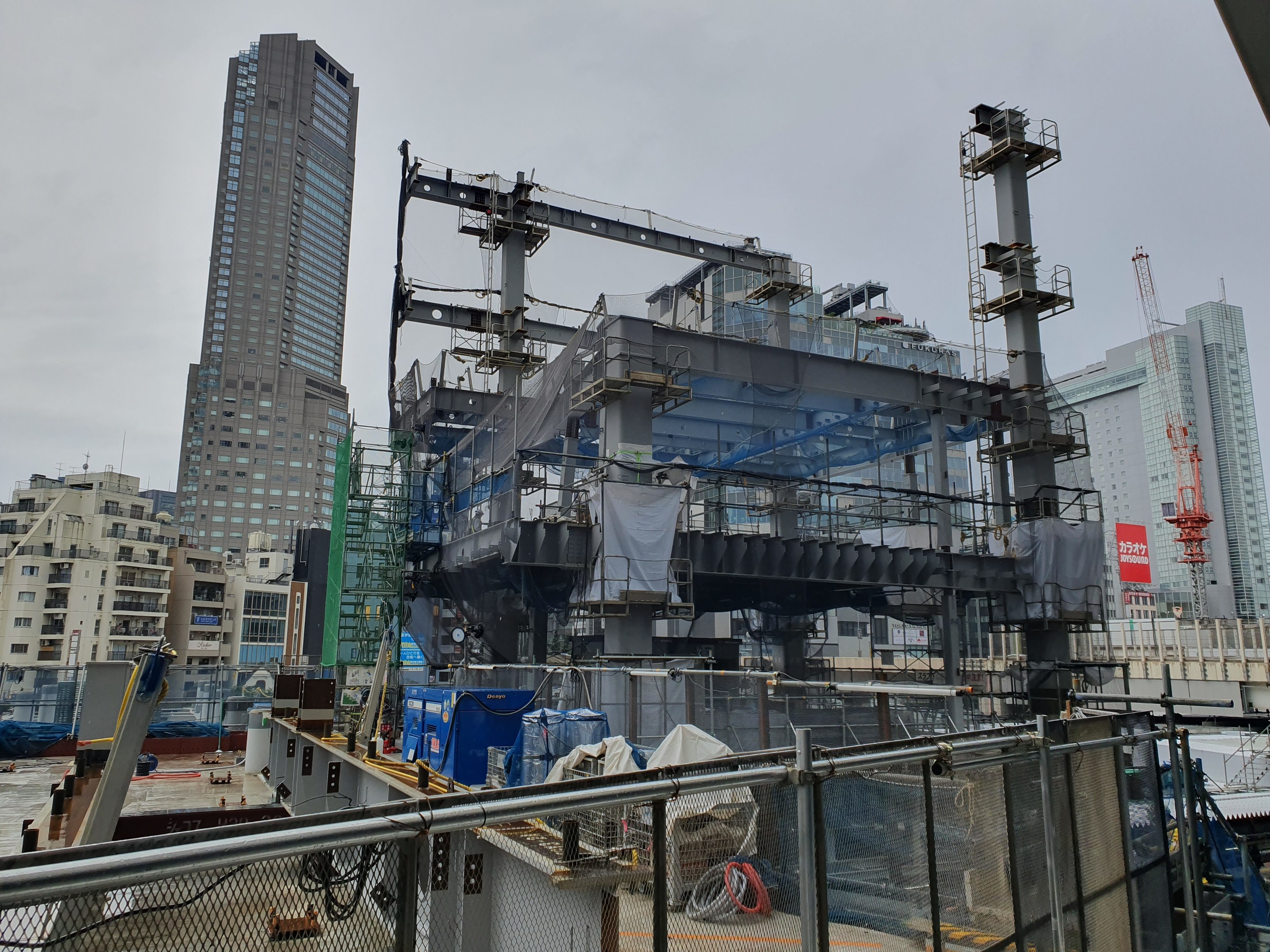 Großbaustelle für ein neues Kraftwerk in einer Stadtumgebung, umgeben von einem Metallzaun, mit Gebäuden, Polen, einer Fahne, Fahrzeugen, Drähten und einem bewölkten Himmel.