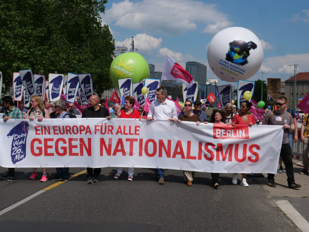 Gruppe von Menschen marschiert bei einer Demonstration gegen Nationalismus in Berlin, trägt ein Banner, Fahnen und Luftballons, mit Bäumen, Gebäuden und einem bewölkten Himmel im Hintergrund.