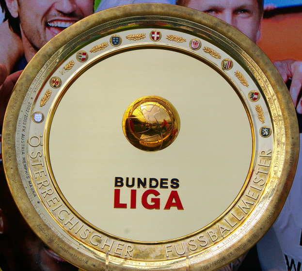 Ein Mann hält ein goldenes Schild mit der Aufschrift "Bundesliga" hoch, im Hintergrund eine Fahne mit drei Personen.