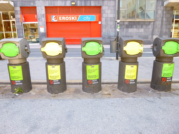Eine Reihe von Parkautomaten entlang einer Straße mit einem Gebäude mit Glasfenstern und einem Rollladen mit Text, sowie einem nahen Müllcontainer.