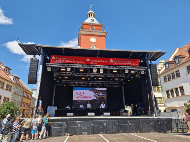 Gruppe von Menschen auf einer Bühne vor einem Uhrenturm beim Kreissparkasse Gotha Musikfestival, mit Gebäuden, Bäumen und einem bewölkten Himmel im Hintergrund.