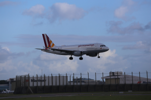 Germanwings Airbus A320-200 beim Abheben vom Frankfurter Airport mit Gras darunter, einem Zaun im Vordergrund, Gebäuden und Bäumen im Hintergrund und einer bewölkten Himmel.
