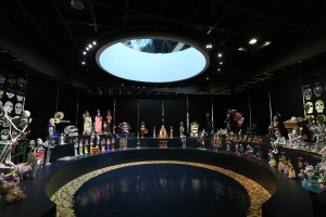 Ein großer Raum voller verschiedener Spielzeuge, darunter Skelette, die auf dem Boden angeordnet sind, mit Schädeln an den Wänden und Lichtern an der Decke, die eine festliche Atmosphäre für das Tag der Toten-Fest schaffen.