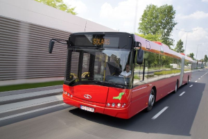 Ein roter Bus fährt auf einer Straße neben einem Gebäude, mit einer Person drinnen, neben einer Wand, Bäumen, Laternenpfählen und einem bewölkten Himmel.