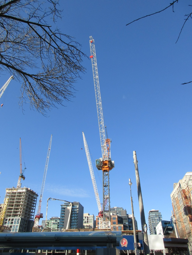 Eine Gruppe von Baukränen auf einer Stadtstraße umgeben von Gebäuden, Bäumen, Pfählen, Brettern und anderen Gegenständen mit dem Himmel im Hintergrund.