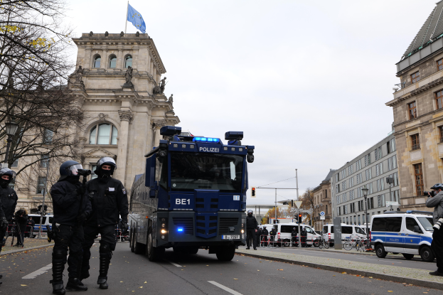 Eine Gruppe von Polizisten in Helmen und mit Waffen in der Hand steht vor einem Gebäude mit Fenstern, Säulen, Bögen und einer Flagge, mit Fahrzeugen auf der Straße, Bäumen, Laternenpfählen und einem bewölkten Himmel im Hintergrund.