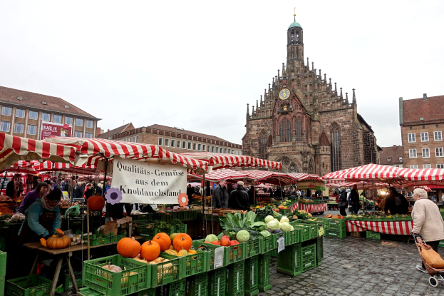 Ein belebter Markt in Nürnberg, Deutschland, mit farbenfrohen Obst- und Gemüsesorten, Menschen mit Taschen und aufgestellten Zelten, sowie Gebäuden und einem Uhrenturm im Hintergrund unter einem sichtbaren Himmel.