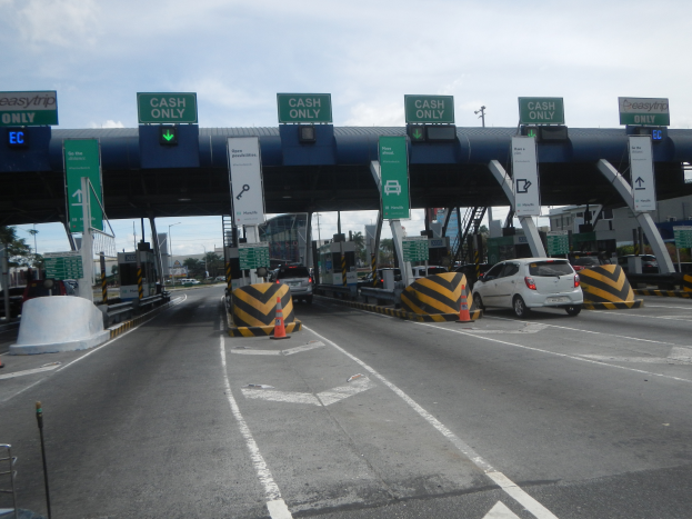 Eine Mautstation an der Seite einer Straße mit darunter fahrenden Autos, umgeben von Verkehrskegeln, Absperrpfählen, Schildern, einem Tor, Laternenmasten, Bäumen, Gebäuden und einem bewölkten Himmel.