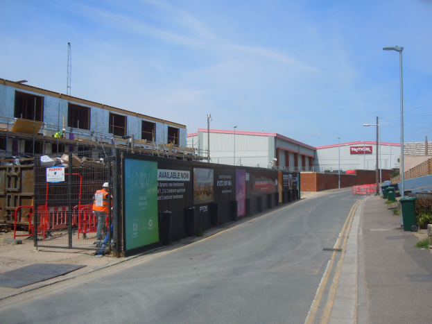 Straße mit einem Gehweg auf der rechten Seite, Laternen, Mülltonnen, Pflanzen und einem Zaun, die zu Gebäuden, einem Turm und dem Himmel führen, mit einer Baustelle und einem unvollendeten Gebäude auf der linken Seite.