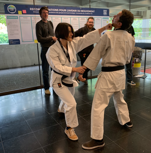 Ein Mann und eine Frau in Karate-Ausrüstung stehen nebeneinander vor einem Plakat, mit einer Glaswand, die Bäume dahinter zeigt, und einem Mülleimer und einem Tisch mit verschiedenen Gegenständen rechts.